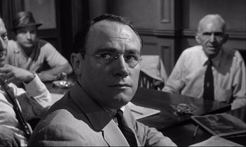 Movie still from “12 Angry Men” (1957), directed by Sidney Lumet – A man with glasses sitting at a table in front of other men; Close Up shot, Over the shoulder angle