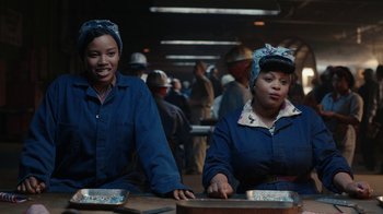 Movie still from “A League of Their Own” (2022), created by Abbi Jacobson – A couple of women sitting at a table in a warehouse; Medium shot, High angle