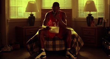 Movie still from “He Got Game” (1998), directed by Spike Lee – A man sitting on top of a bed eating a piece of food; Medium shot, Low angle