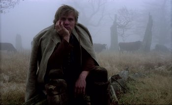 Movie still from “Heart of Glass” (1976), directed by Werner Herzog – A man sitting on the ground in a field with cows; Medium shot, Low angle
