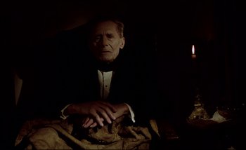 Movie still from “Heart of Glass” (1976), directed by Werner Herzog – An old man sitting in a dark room with a candle in the background; Medium shot, High angle