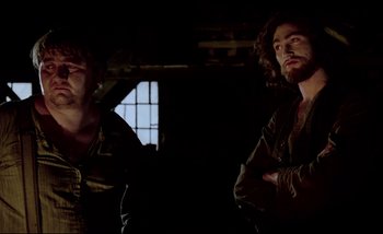 Movie still from “Heart of Glass” (1976), directed by Werner Herzog – Two men standing next to each other in a dark room; Medium shot, Low angle