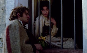 Movie still from “Heart of Glass” (1976), directed by Werner Herzog – A man and a woman sitting next to each other in front of bars; Medium shot, Low angle