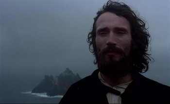 Movie still from “Heart of Glass” (1976), directed by Werner Herzog – A man with long hair and a goatee standing in front of a body of water; Close Up shot, Low angle
