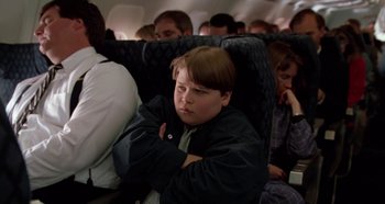 Movie still from “Heavyweights” (1995), directed by Steven Brill – A young boy sitting on an airplane with his arms crossed; Medium shot, High angle