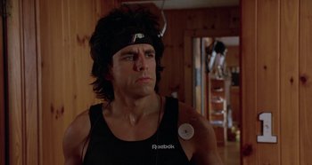 Movie still from “Heavyweights” (1995), directed by Steven Brill – A man in a black tank top with a button on his head; Close Up shot, Low angle