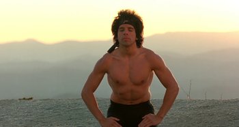 Movie still from “Heavyweights” (1995), directed by Steven Brill – A shirtless man with long hair is standing in front of a body of water; Medium shot, Low angle