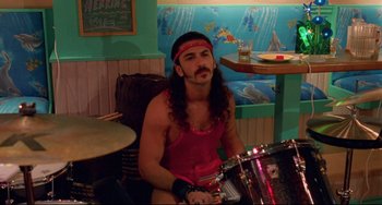 Movie still from “Hedwig and the Angry Inch” (2001), directed by John Cameron Mitchell – A man with long curly hair and a headband is sitting in front of a drum; Medium shot, High angle