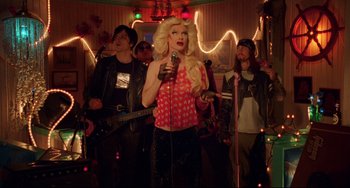 Movie still from “Hedwig and the Angry Inch” (2001), directed by John Cameron Mitchell – A group of people standing in a room with a microphone; Medium shot, Over the shoulder angle