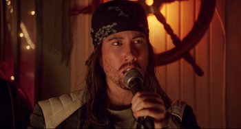 Movie still from “Hedwig and the Angry Inch” (2001), directed by John Cameron Mitchell – A man with long brown hair wearing a black hat and holding a microphone in front of him; Close Up shot, Over the shoulder angle