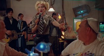 Movie still from “Hedwig and the Angry Inch” (2001), directed by John Cameron Mitchell – A woman is singing into a microphone at a party; Medium shot, Over the shoulder angle
