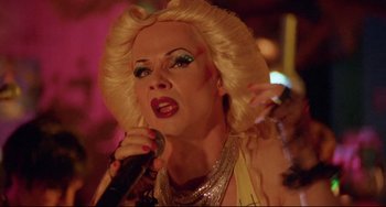 Movie still from “Hedwig and the Angry Inch” (2001), directed by John Cameron Mitchell – A woman with blonde hair and a silver necklace holding a microphone in front of her face; Close Up shot, Low angle