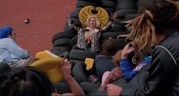 Movie still from “Hedwig and the Angry Inch” (2001), directed by John Cameron Mitchell – A woman sitting on a pile of tires with people sitting around her; Wide shot, Low angle