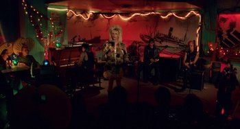 Movie still from “Hedwig and the Angry Inch” (2001), directed by John Cameron Mitchell – A woman is singing into a microphone in front of a group of people; Wide shot, High angle