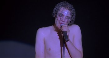 Movie still from “Hedwig and the Angry Inch” (2001), directed by John Cameron Mitchell – A man with a cross on his head is holding a microphone; Close Up shot, Low angle