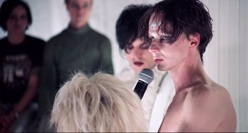 Movie still from “Hedwig and the Angry Inch” (2001), directed by John Cameron Mitchell – A person with a microphone in front of a group of people; Close Up shot, Over the shoulder angle