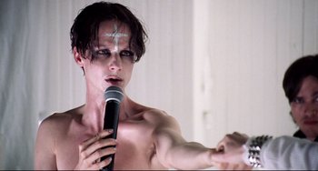 Movie still from “Hedwig and the Angry Inch” (2001), directed by John Cameron Mitchell – A shirtless man holding a microphone in front of a wall; Close Up shot, Low angle