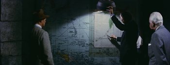 Movie still from “Hell and High Water” (1954), directed by Samuel Fuller – A person holding a hat and a map in a room; Medium shot, High angle