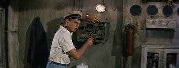 Movie still from “Hell and High Water” (1954), directed by Samuel Fuller – A man in a hat is holding an old radio; Medium shot, High angle
