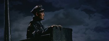 Movie still from “Hell and High Water” (1954), directed by Samuel Fuller – A man in a black jacket and a hat is standing in front of a dark sky; Medium shot, Low angle