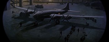 Movie still from “Hell and High Water” (1954), directed by Samuel Fuller – An airplane is parked in a hangar at night with people walking around; Extreme Wide shot, High angle