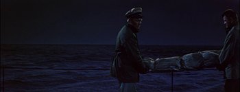 Movie still from “Hell and High Water” (1954), directed by Samuel Fuller – A man in a cap and jacket standing in front of a body of water at night; Wide shot, High angle
