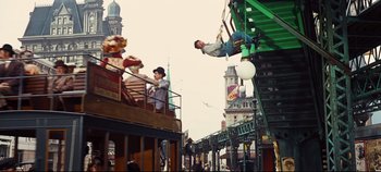 Movie still from “Hello, Dolly!” (1969), directed by Gene Kelly – A man hanging from a metal structure on a street; Wide shot, Low angle