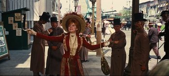 Movie still from “Hello, Dolly!” (1969), directed by Gene Kelly – A woman in a red dress and a straw hat is on a street; Medium shot, Over the shoulder angle