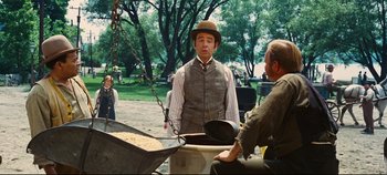 Movie still from “Hello, Dolly!” (1969), directed by Gene Kelly – A man in a hat standing in front of a pan of food; Medium shot, Over the shoulder angle