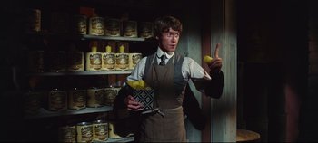 Movie still from “Hello, Dolly!” (1969), directed by Gene Kelly – A man holding a tray of lemons in front of shelves of cans; Medium shot, Over the shoulder angle