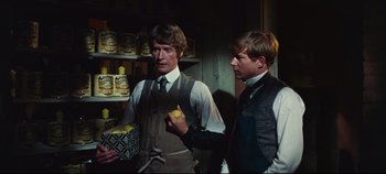 Movie still from “Hello, Dolly!” (1969), directed by Gene Kelly – Two young men standing next to each other holding an apple; Medium shot, Over the shoulder angle