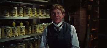 Movie still from “Hello, Dolly!” (1969), directed by Gene Kelly – A man in a vest and tie standing in front of shelves of cans; Medium shot, Over the shoulder angle