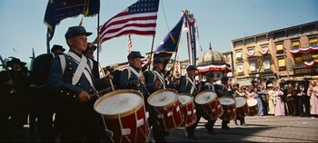 Movie still from “Hello, Dolly!” (1969), directed by Gene Kelly – A group of men in uniform marching down a street with drums; Wide shot, Low angle