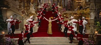 Movie still from “Hello, Dolly!” (1969), directed by Gene Kelly – A woman in a gold dress standing on top of a red staircase; Extreme Wide shot, High angle
