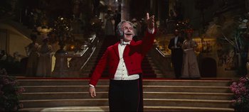 Movie still from “Hello, Dolly!” (1969), directed by Gene Kelly – A man wearing a red jacket and a white bow tie standing in front of a staircase; Medium shot, Low angle