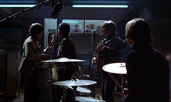 Movie still from “Help!” (1965), directed by Richard Lester – A group of people that are standing around a room; Wide shot, Over the shoulder angle