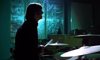 Movie still from “Help!” (1965), directed by Richard Lester – A man smoking a cigarette while playing drums in a room; Medium shot, Over the shoulder angle