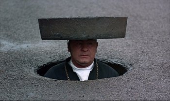 Movie still from “Help!” (1965), directed by Richard Lester – A man with a hat on his head looking into a hole in the ground; Wide shot, Overhead angle