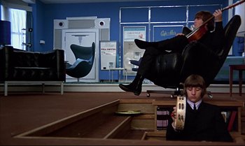 Movie still from “Help!” (1965), directed by Richard Lester – A man sitting on steps in front of a black chair; Wide shot, Low angle