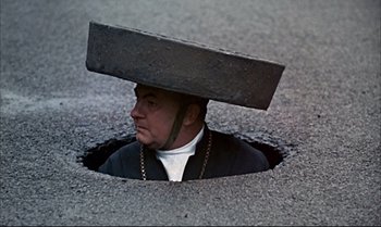 Movie still from “Help!” (1965), directed by Richard Lester – A man with a large brick on his head is in a hole in the ground; Wide shot, Overhead angle