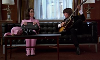 Movie still from “Help!” (1965), directed by Richard Lester – A woman in a pink suit sitting next to a man in a black suit; Wide shot, Low angle