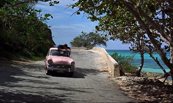 Movie still from “Help!” (1965), directed by Richard Lester – An old pink car driving down a road next to the ocean; Wide shot, High angle