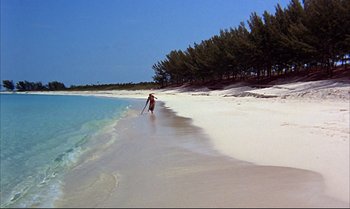 Movie still from “Help!” (1965), directed by Richard Lester – A person walking on the beach with a surfboard; Extreme Wide shot, High angle