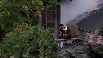 Movie still from “Henry & June” (1990), directed by Philip Kaufman – A man sitting on a chair on a porch with a guitar; Wide shot, High angle