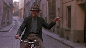 Movie still from “Henry & June” (1990), directed by Philip Kaufman – A man riding a bike down the street wearing a hat; Medium shot, Over the shoulder angle