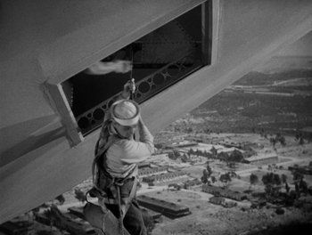 Movie still from “Here Comes the Navy” (1934), directed by Lloyd Bacon – A man hanging from the side of a building on a rope; Wide shot, Low angle