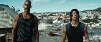 Movie still from “Hereafter” (2010), directed by Clint Eastwood – Two men standing on the roof of a building looking at a man laying on the ground in the water; Medium shot, High angle
