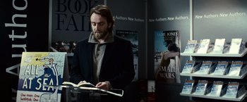 Movie still from “Hereafter” (2010), directed by Clint Eastwood – A man standing in front of a table reading a book; Medium shot, Over the shoulder angle