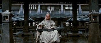 Movie still from “Hero” (2002), directed by Yimou Zhang – An old man sitting on a bench in front of a building; Wide shot, Low angle