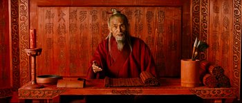 Movie still from “Hero” (2002), directed by Yimou Zhang – An old man sitting in front of a wooden wall; Medium shot, Low angle